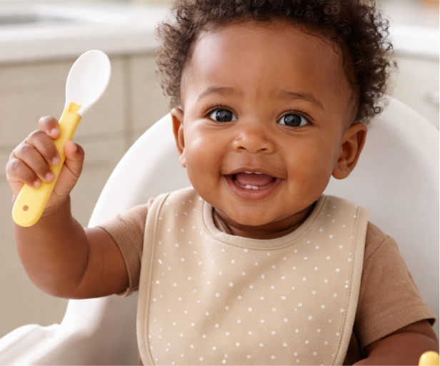 Baby with curly hair holding a yellow spoon and smiling
